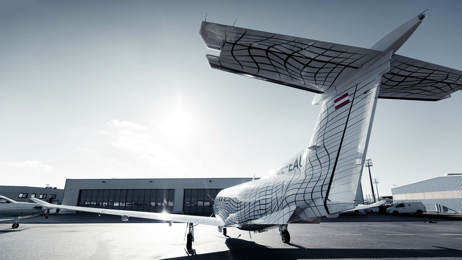 Ein weißes Flugzeug von Pilart Air mit einem Gittermuster auf dem Rumpf steht unter klarem Himmel auf dem Rollfeld eines Flughafens, im Hintergrund sind ein Hangar und andere Flugzeuge zu sehen.