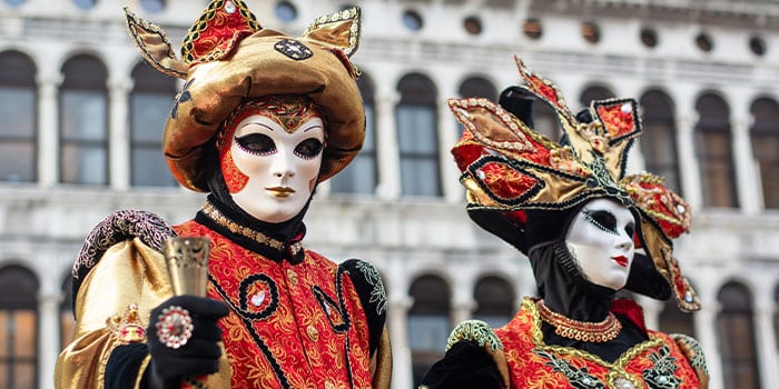 Zwei Personen in aufwendigen venezianischen Kostümen und Masken stehen vor einem historischen Gebäude mit Bogenfenstern.