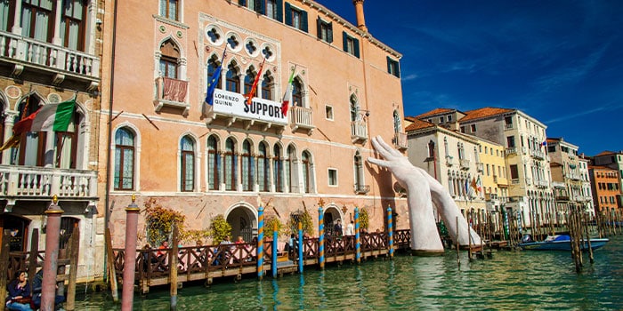 Große Skulptur von Händen, die aus dem Wasser ragen und ein Gebäude in Venedig stützen. Farbenfrohe Gebäude und klarer blauer Himmel im Hintergrund.