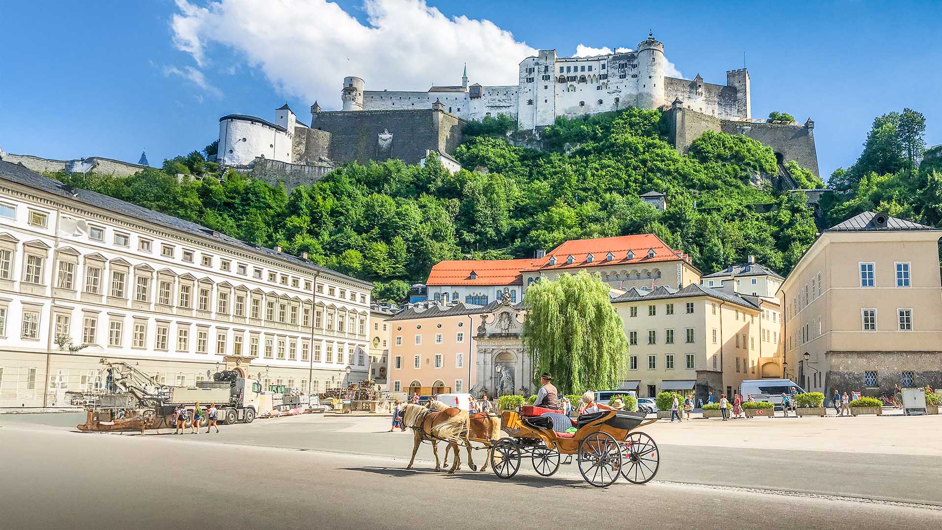 Eine Pferdekutsche auf einem sonnigen Platz mit Gebäuden und einer großen Festung auf einem Hügel im Hintergrund.