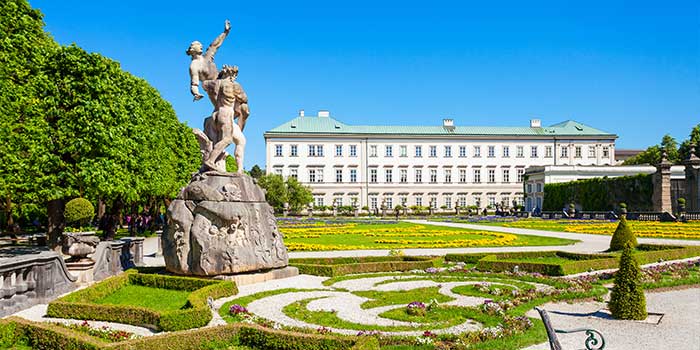 Barocke Skulpturen und gepflegte Gärten vor einem großen historischen Gebäude unter einem klaren blauen Himmel. Schloss Mirabell.