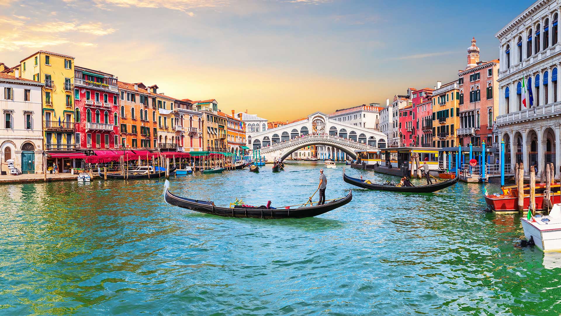 Gondeln auf dem Canale Grande in Venedig bei Sonnenuntergang, umgeben von farbenfrohen Gebäuden und der Rialtobrücke im Hintergrund.