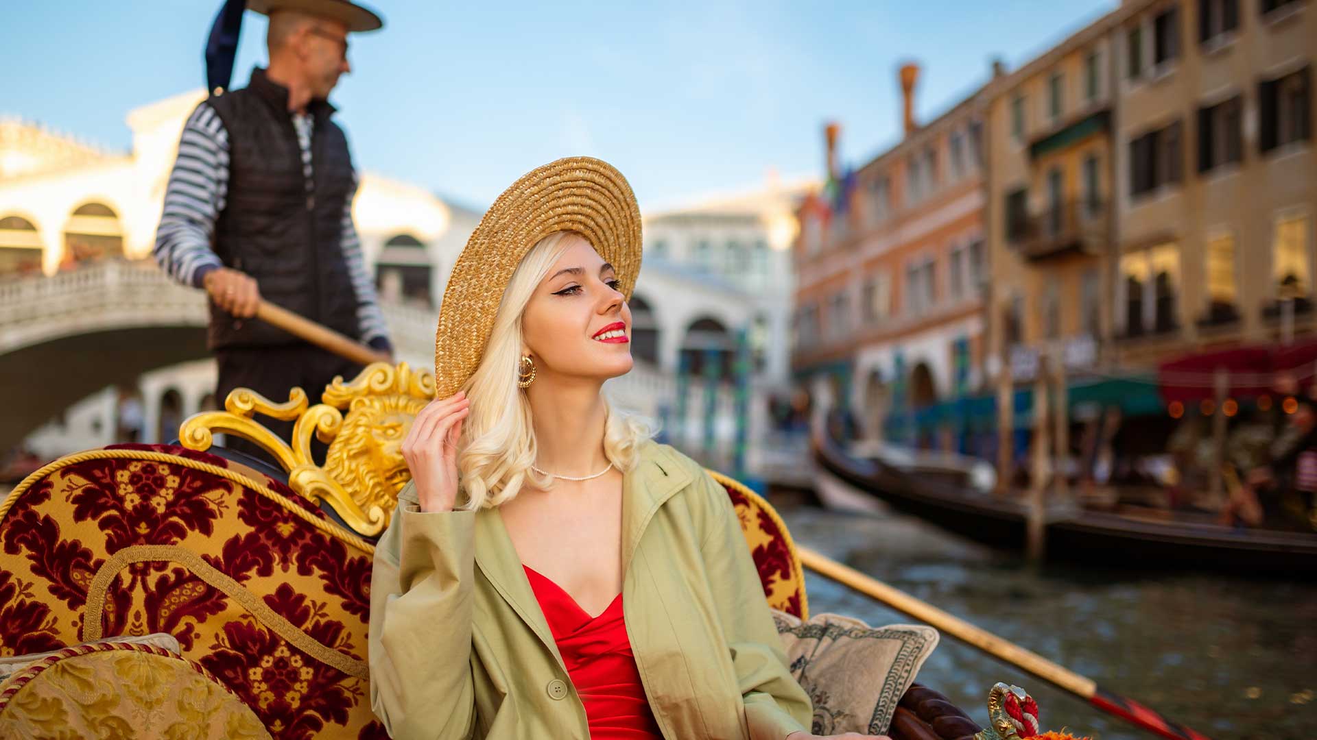 Eine Frau mit Strohhut und rotem Kleid sitzt auf einer geschmückten Gondel, während ein Gondoliere durch einen Kanal in Venedig steuert. Im Hintergrund sind Gebäude und eine Brücke zu sehen.