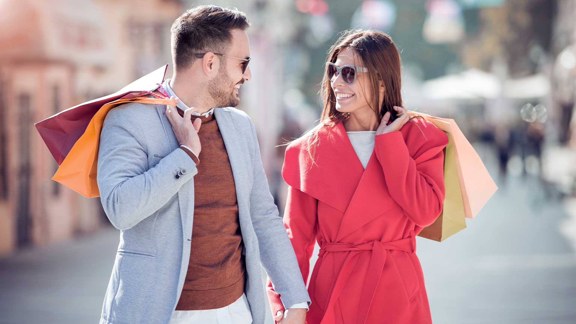 Ein Mann und eine Frau gehen mit Einkaufstüten eine Straße entlang und lächeln sich an. Beide tragen Sonnenbrillen.
