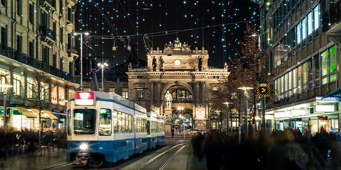 Nachtszene einer belebten Straße mit einer fahrenden blauen Straßenbahn, darüber hängenden Weihnachtslichtern und einem historischen Gebäude im Hintergrund.