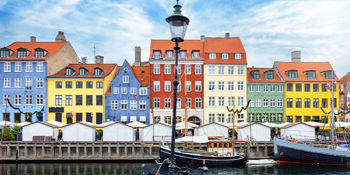 Farbenfrohe Gebäude säumen die Uferpromenade im Kopenhagener Nyhavn unter einem bewölkten Himmel. Im Vordergrund sind Boote zu sehen, die vor Anker liegen.