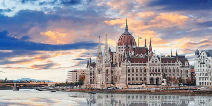 Das ungarische Parlamentsgebäude spiegelt sich bei Sonnenuntergang in der Donau, darüber ein teilweise bewölkter Himmel.