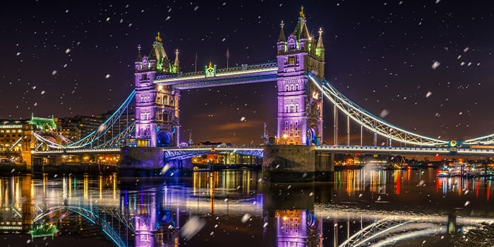 Die Tower Bridge ist nachts violett und grün beleuchtet, während fallender Schnee sich im Fluss darunter spiegelt.