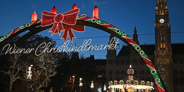 Beleuchteter Bogen mit Schleife am Eingang des Wiener Christkindlmarkts, mit festlichen Lichtern und einem Uhrturm im Hintergrund.