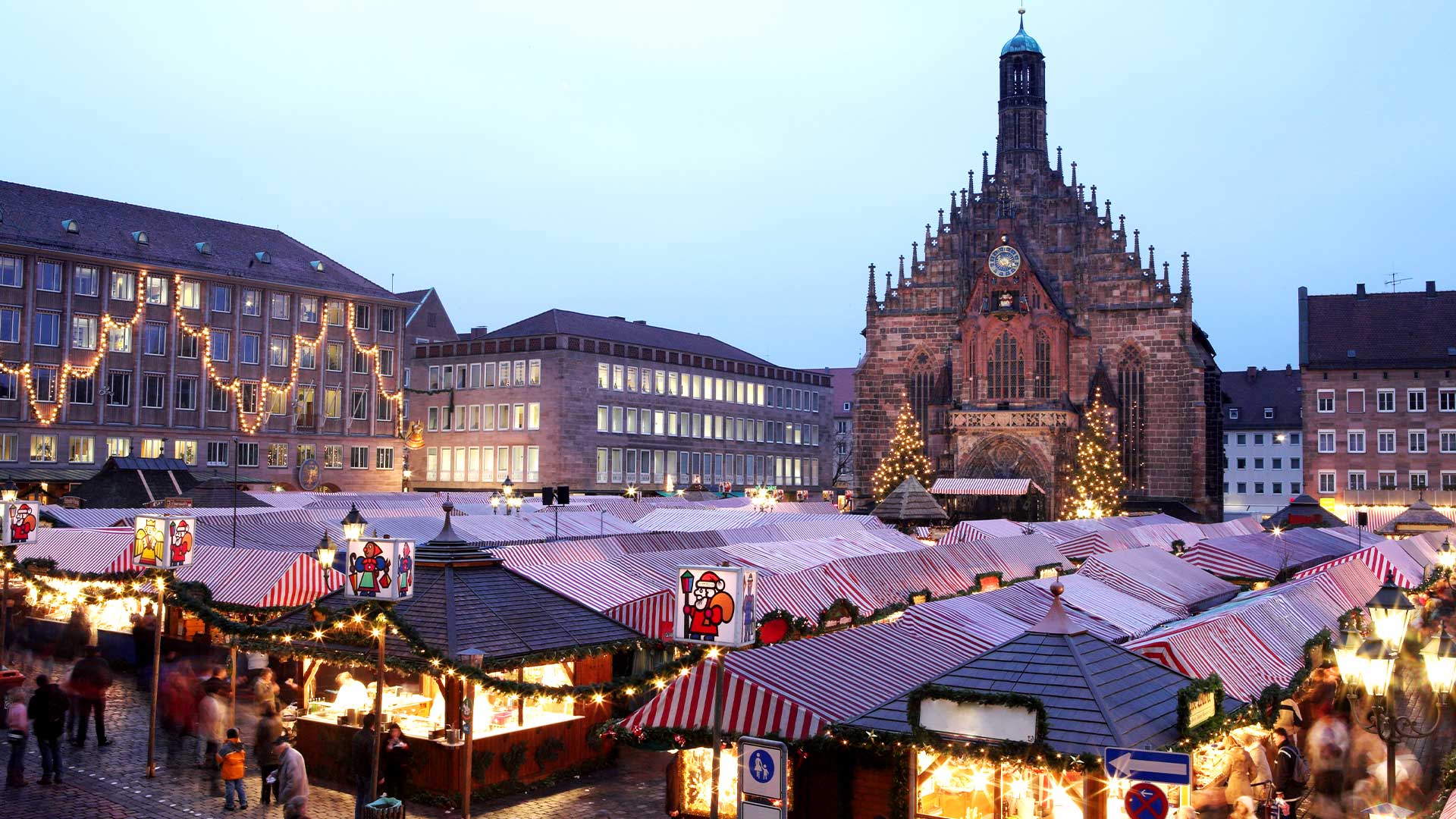 Luftaufnahme eines geschäftigen Weihnachtsmarkts mit gestreiften Zelten und festlichen Lichtern vor einer großen, historischen Kirche in der Abenddämmerung.