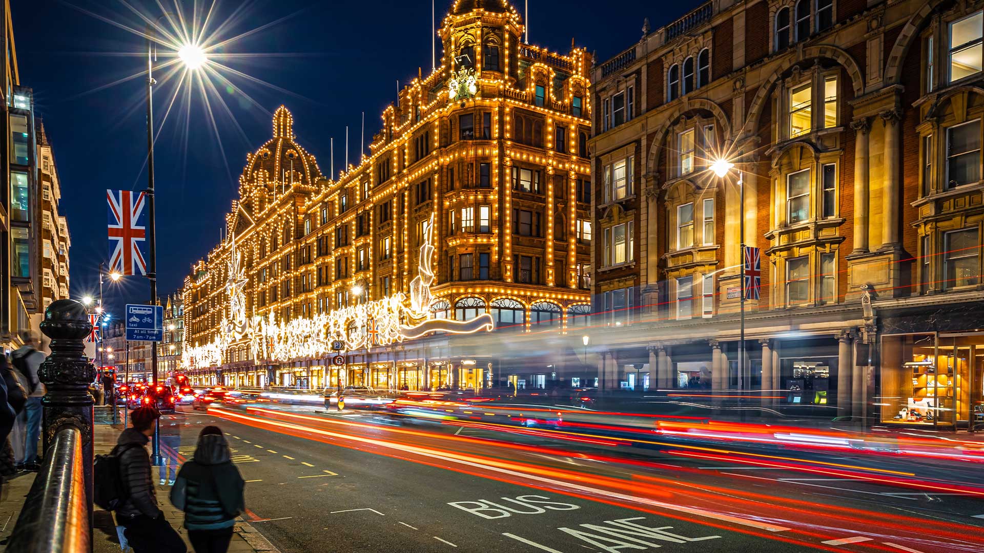 Eine belebte Londoner Straße bei Nacht mit Lichtspuren von Fahrzeugen und einem hell erleuchteten, reich verzierten Gebäude.