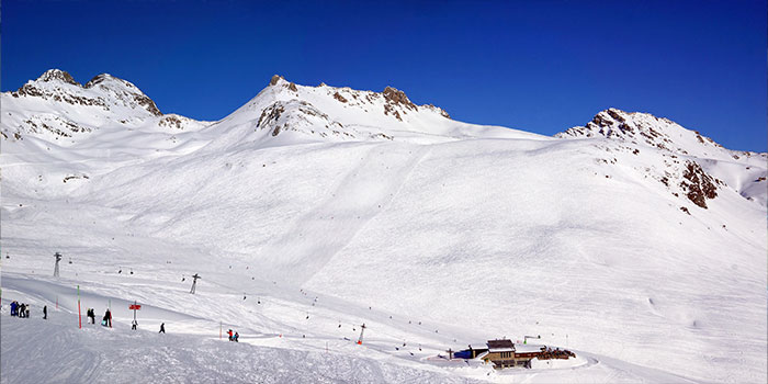 Schneebedeckte Berge mit klarem, blauem Himmel; Skifahrer und Snowboarder auf einem Hang in Richtung eines kleinen Gebäudes.
