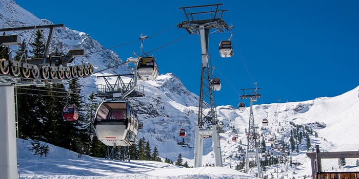 An einem klaren Tag transportiert eine Skigondel Menschen auf einen schneebedeckten Berg, im Hintergrund sind Bäume und blauer Himmel zu sehen.