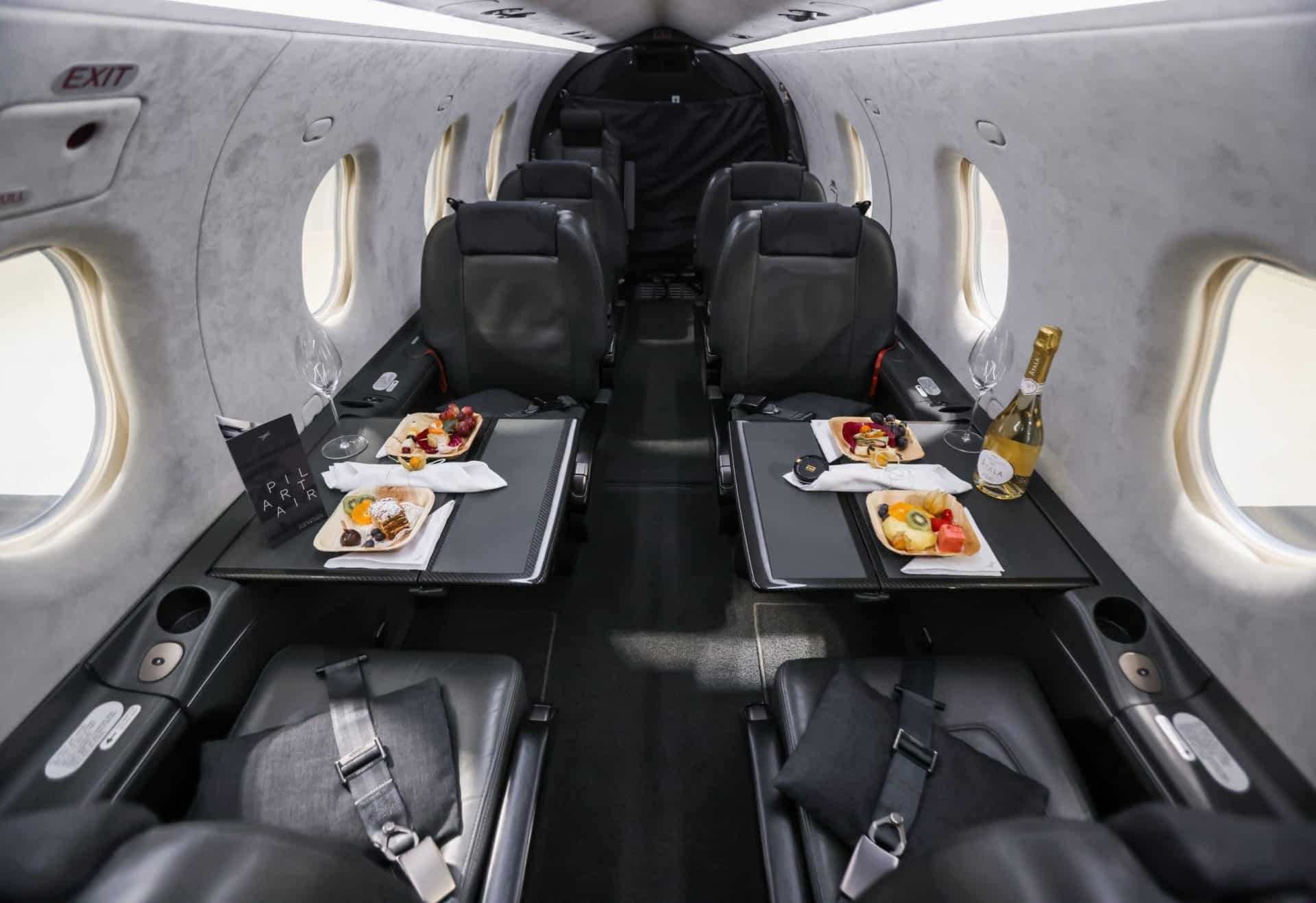 Innenansicht einer PC12 von Pilart Air mit zwei für eine Mahlzeit gedeckten Tischen. Auf jedem Tisch stehen Teller mit Essen und eine Flasche Champagner, umgeben von Ledersitzen.