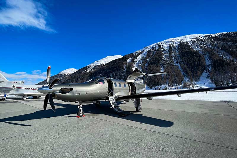 Ein kleines Privatflugzeug mit offener Tür steht auf einem Rollfeld, im Hintergrund sind schneebedeckte Berge unter einem klaren blauen Himmel zu sehen.