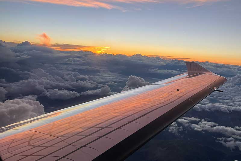 Blick aus einem Flugzeugflügel bei Sonnenuntergang, mit Wolken unten und einem bunten Himmel darüber.