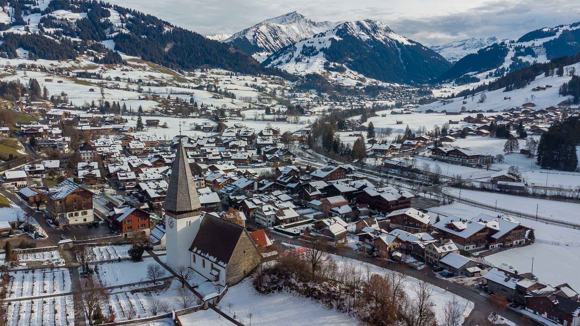 Luftaufnahme eines verschneiten Dorfes mit einer Kirche, umgeben von Bergen.