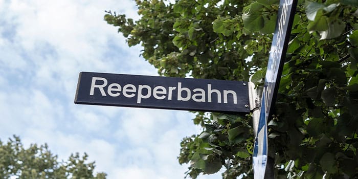 Straßenschild „Reeperbahn“ mit Bäumen im Hintergrund unter einem bewölkten Himmel.
