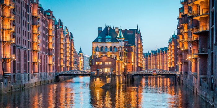 Historisches Lagerhausviertel in der Abenddämmerung mit beleuchteten Gebäuden entlang eines Kanals und einem zentralen, durch Brücken verbundenen Gebäude, das sich im Wasser spiegelt.