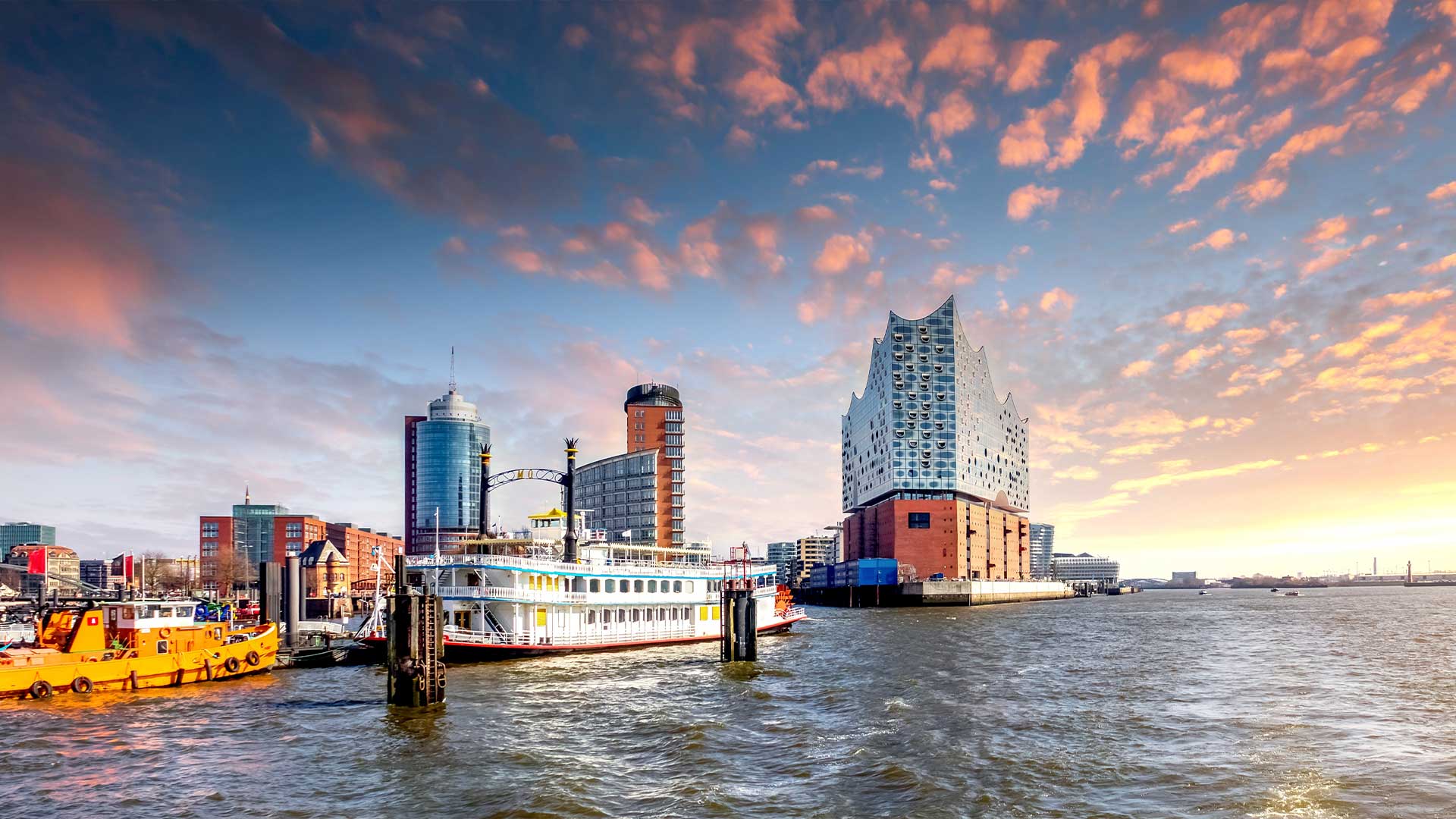 Eine am Wasser gelegene Stadtlandschaft mit einem modernen Konzertsaal, einem angedockten Schiff und Gebäuden unter einem leuchtenden Sonnenuntergangshimmel.