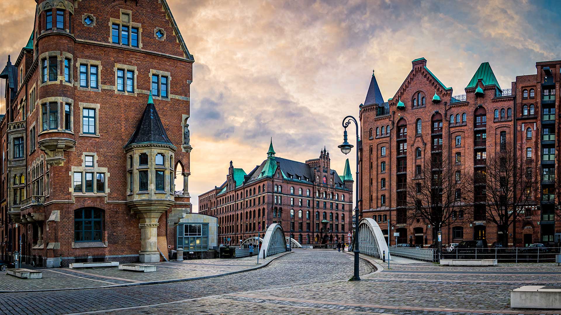 Historische Backsteingebäude umgeben eine Kopfsteinpflasterstraße mit Bogenbrücken und einem Laternenpfahl unter einem bewölkten Himmel.