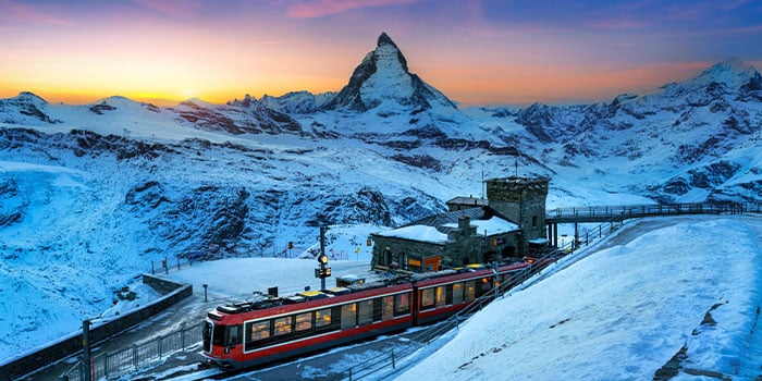 Ein roter Zug fährt bei Sonnenuntergang durch eine verschneite Bergstation, im Hintergrund ist das Matterhorn zu sehen.