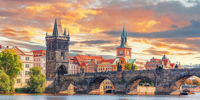 Historische Steinbrücke mit Türmen und einer lebendigen Stadtlandschaft unter einem farbenfrohen Himmel bei Sonnenuntergang.