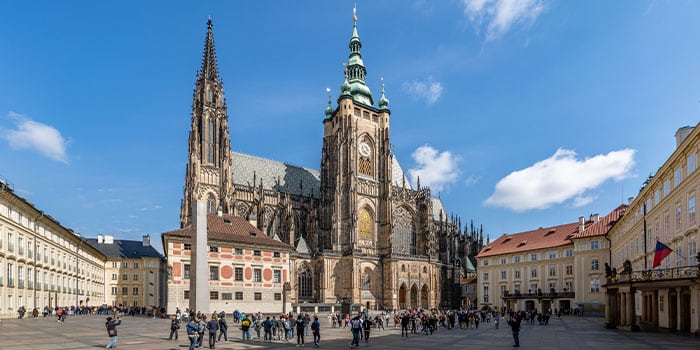 Eine Menschenmenge steht vor einer großen, reich verzierten Kathedrale mit einem Glockenturm, umgeben von anderen historischen Gebäuden unter einem klaren blauen Himmel.